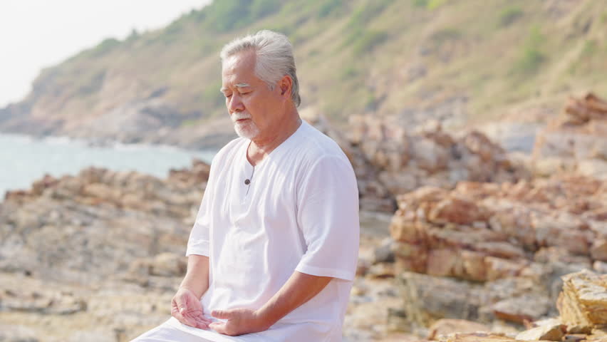 Asian senior man practicing meditation with ocean nature on rocky coastal hill at summer sunset. Retirement elderly people do outdoor relaxing yoga exercise. Mental health care and motivation concept.