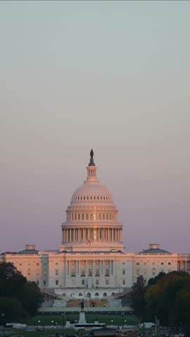 Vertical Video The United States Capitol Building stands tall against a soft pink sunset sky, with a construction crane visible. Trees line the front of the building, and a few people walk 
