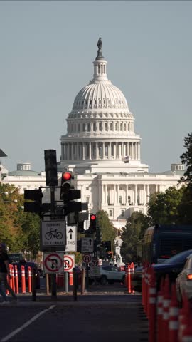 Vertical Video street scene in Washington DC with the Capitol Building in the background. Traffic is stopped at a red light and pedestrians cross the street. Several signs, trees, and streetlights