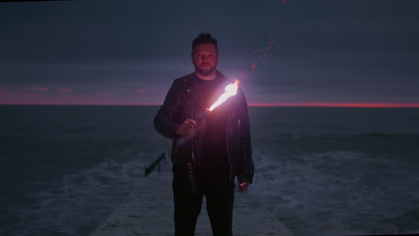 Portrait: a man traveler in a leather jacket holds a red signal light in his hands. Male stands on the breakwater, burning a fire stick or torch. Sea or ocean in the evening, big wave. Slow motion