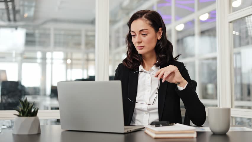 Businesswoman working on a laptop in the office. Woman focused on her work, holding her glasses while reviewing data or emails on her computer.