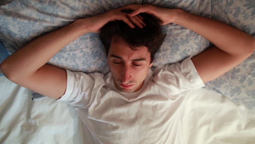 Man lays down on a bed, looking off to the side with a thoughtful expression. His hands are on his head, suggesting a moment of contemplation or concern in a calm, domestic setting