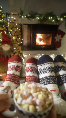 Couple enjoying cocoa with marshmallows by warm fireplace and warming up their feet in woolen socks with Christmas ornaments and Christmas tree and decorations background. Cozy Winter, Christmas and