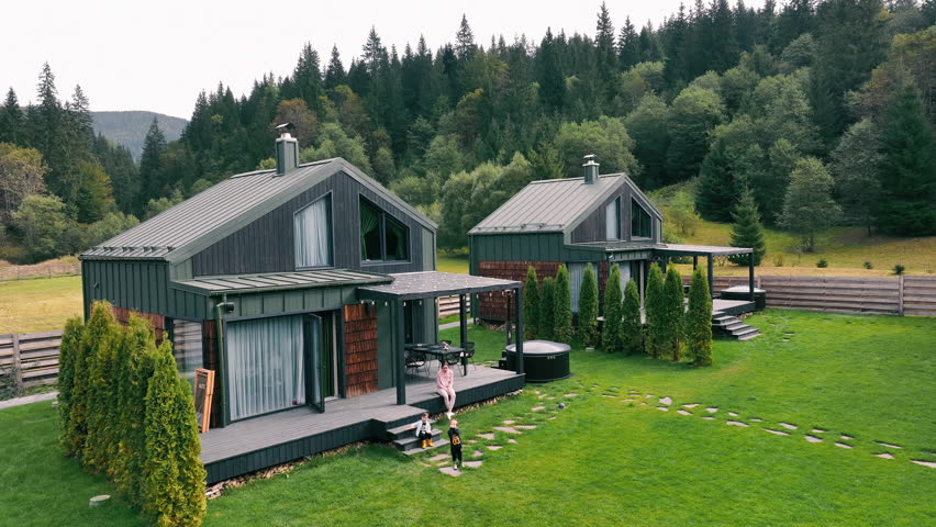 Modern wooden houses for recreation in the mountains. A mother with two small children near the house. Two modern cabins with dark siding and wooden