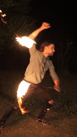 Vertical video. A young man conducts a fire show at night. In each hand he has a chain with a flaming tip. He waves them in the air. Dressed in a gray shirt and black pants, he expertly maneuvers fire