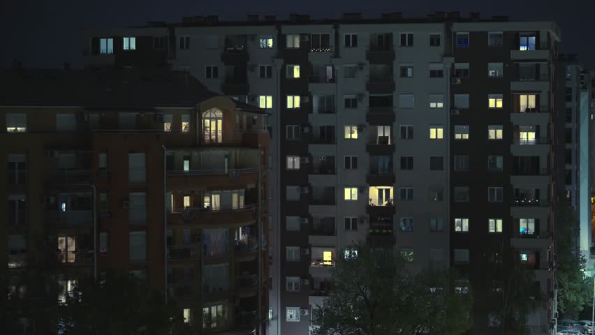Establishing shot of apartments with lit windows at night in an urban setting with a street and green trees