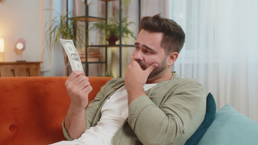 Poor Caucasian young man upset over insufficient amount of money, counting dollar banknotes. Financial crisis. Bankruptcy. Poverty and destitution. Upset adult guy sitting on sofa couch at home.