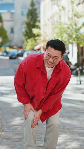 Sick ill Asian middle-aged man suffer from painful discomfort severe knee joint ache, leg muscles, could not walk outdoors. Chinese guy massaging knees illness cramps rheumatism on urban city street