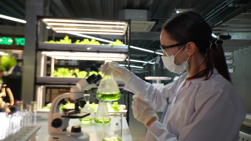 A scientist wearing a mask and gloves works in a lab, examining a green liquid in a beaker and using a microscope. The lab setup is well-lit, reflecting a focus on research, science, and experimentati - Powered by Shutterstock - Get 15% off with code: PIKWIZARD15