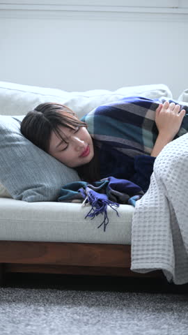 Close-up vertical video of a Japanese woman lying on a sofa, sick with a cold. Shivering with cold.