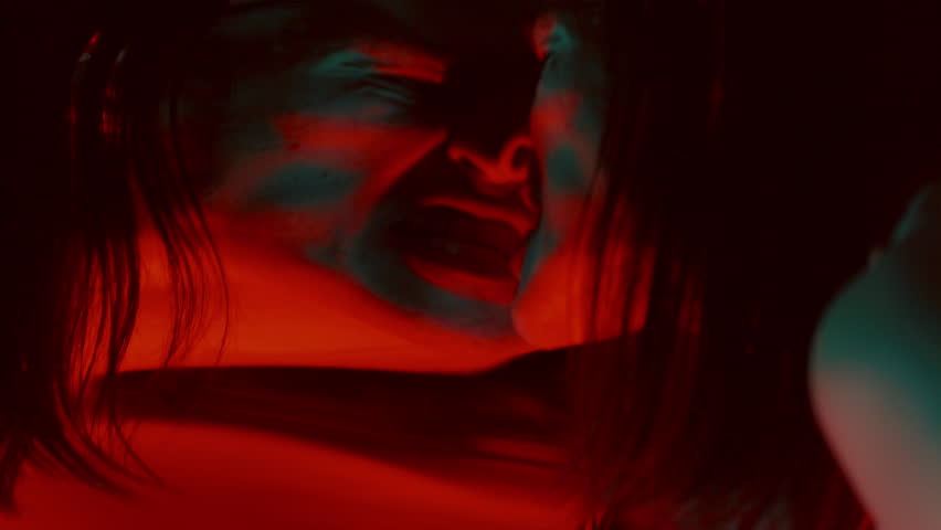 Creepy man pressing his face against distorting mirror, screaming madly with frightening expression in dark studio with red lighting