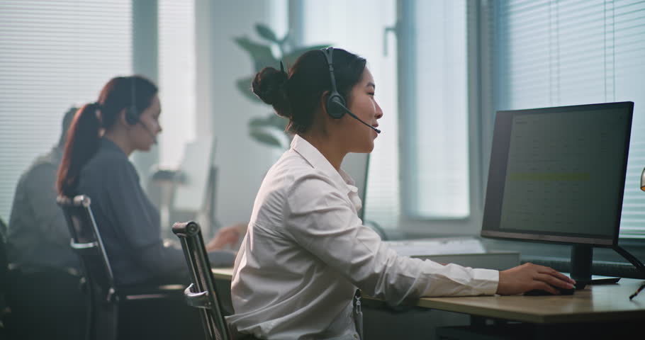 Multiethnic team of technical support operators working in modern call center office. Asian female helpdesk specialist in headset consults client via call, uses computer, providing customer service.