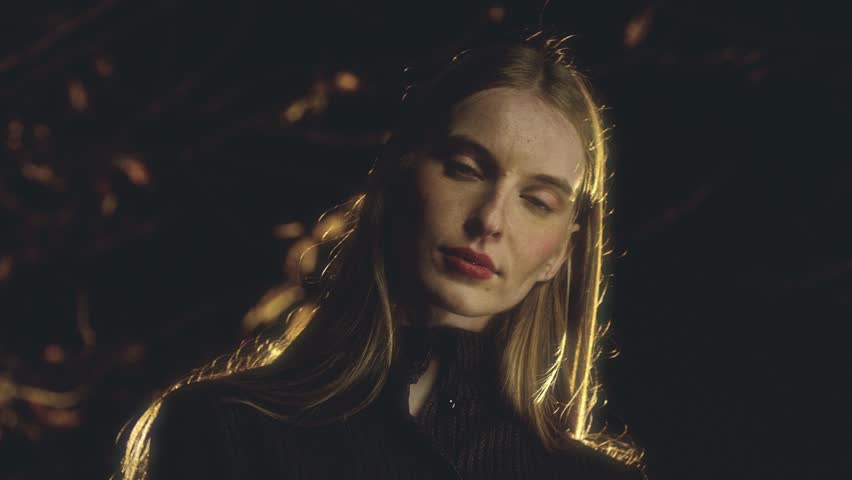 Woman with long blonde hair and a black turtleneck is posing in front of a blurred golden background. She opens and closes her eyes, showcasing her makeup