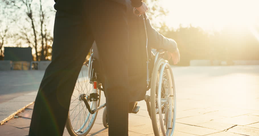 Hispanic man taking care of old disabled friend in wheelchair. Friends spending time together in autumn park. Healthcare, disability, mobility, friendship. Close up 4k shot.