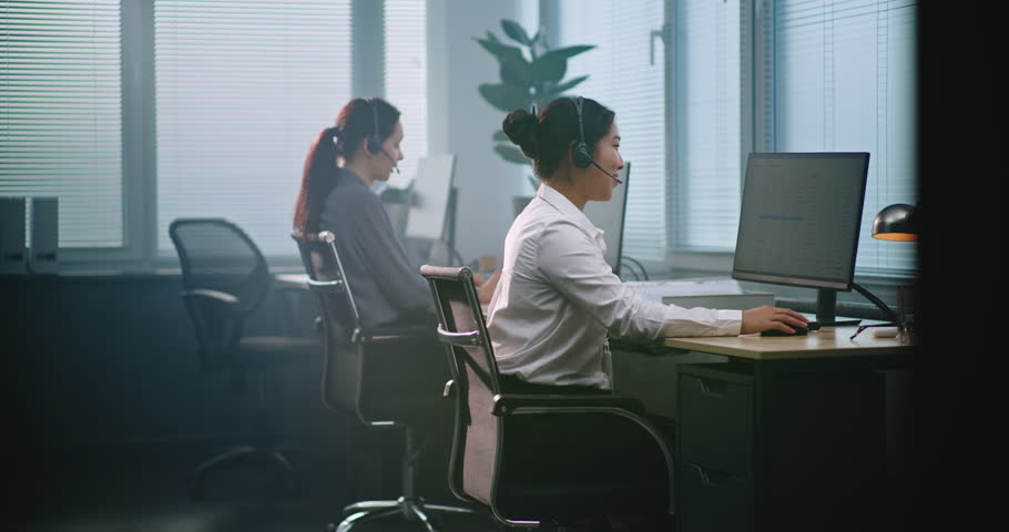 Multiethnic team of technical support operators working in modern call center office. Asian female helpdesk specialist talks on headset, uses computer while providing online tech assistance for client