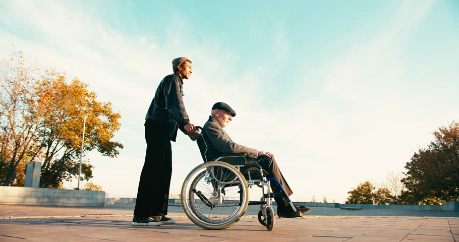 Hispanic man taking care of old disabled friend in wheelchair. Friends spending time together in autumn park. Healthcare, disability, mobility, friendship.