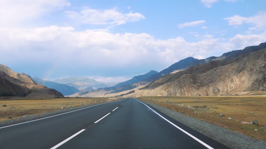 Driving on empty asphalt road winds through mountain valley, surrounded by yellow grass in prairie under sunny sky filled with clouds. View from vehicle driving along the highway. Road trip in summer