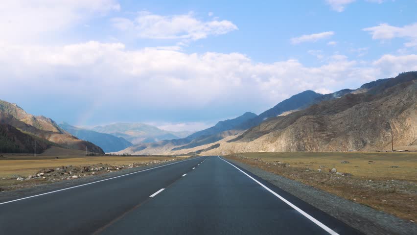 Driving on empty asphalt road winds through mountain valley, surrounded by yellow grass in prairie under sunny sky filled with clouds. View from vehicle driving along the highway. Road trip in summer