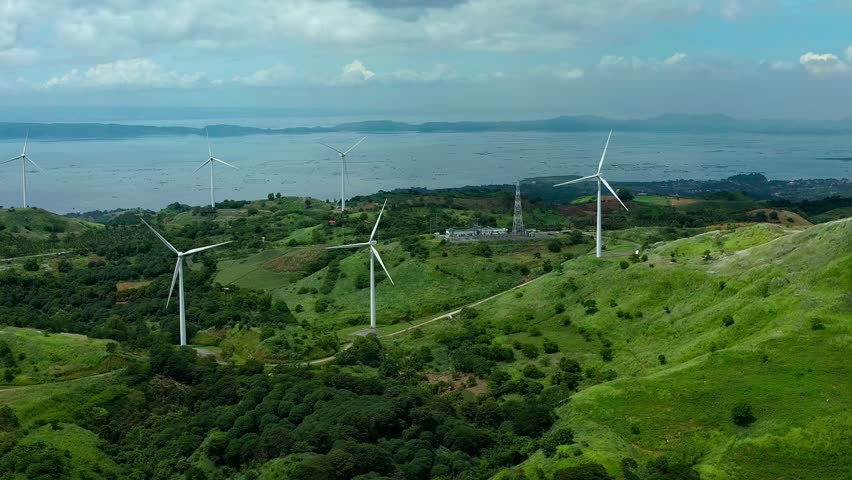 Aerial view of street on green hills with installed wind mills. Sea with islands during cloudy day in Philippines, Rizal Pililla. Wide shot.