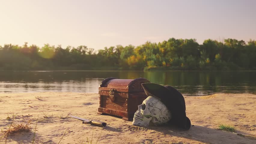 Old style brown chest stands on beach sand sea with pirate skull in a cocked hat, sword. Pirates Treasure chest wooden box with a lock. Shot close-up object fantasy game. Nature summer sun light flare