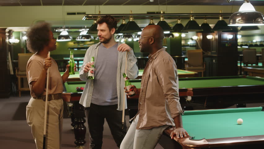 Full medium shot of diverse female and male friends gathered together in club for billiard game and drinking beer