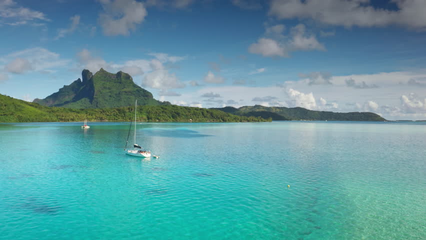 Two sailboats yachts anchored in turquoise waters coast of Bora Bora lagoon, green mountains rising under bright blue sky. Remote wild nature paradise, exotic summer luxury travel. Drone aerial flight
