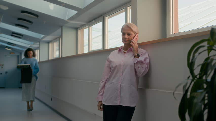Medium shot of young Biracial female designer assistant showing textile samples to mature Caucasian busy brand CEO woman having phone call in modern office hallway