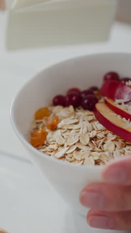 Oats are being topped with a drizzle of milk, enhancing the colorful mix of fruits like apples and berries in a bowl on a bright kitchen countertop