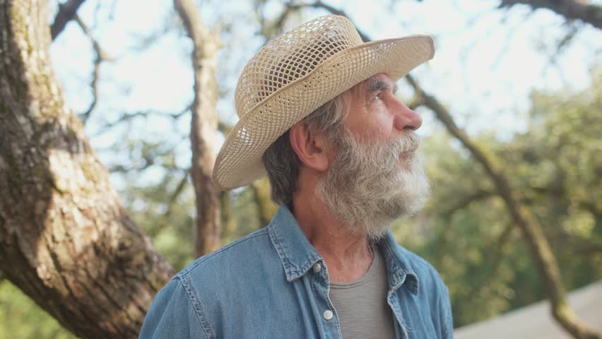 Close facial portrait of older Caucasian male with long grey beard. Experienced farmer looking at camera. Man spending time in old garden. Wearing yellow hat and jeans shirt. Smiling.