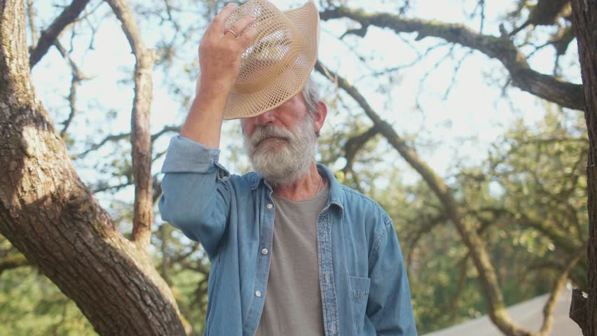 View from below of handsome Caucasian man putting on his yellow hat. Male with blue shirt and grey beard. Experienced farmer spending his free time in woods. Checking health of plants.
