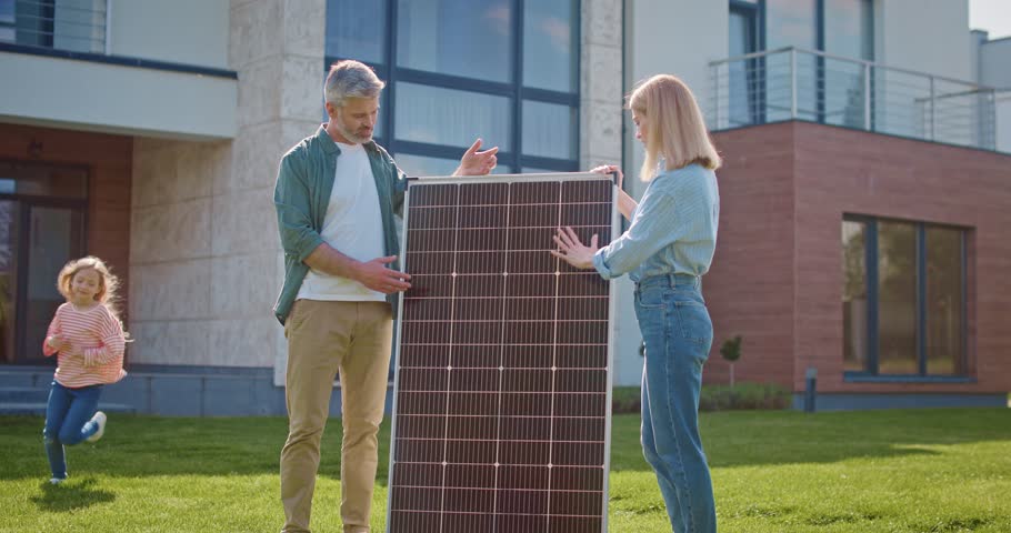 Lovely Caucasian family standing outside of their house. Husband and wife discussing about solar panel. Kids running around them while playing. Man explaining to his wife how solar energy works.