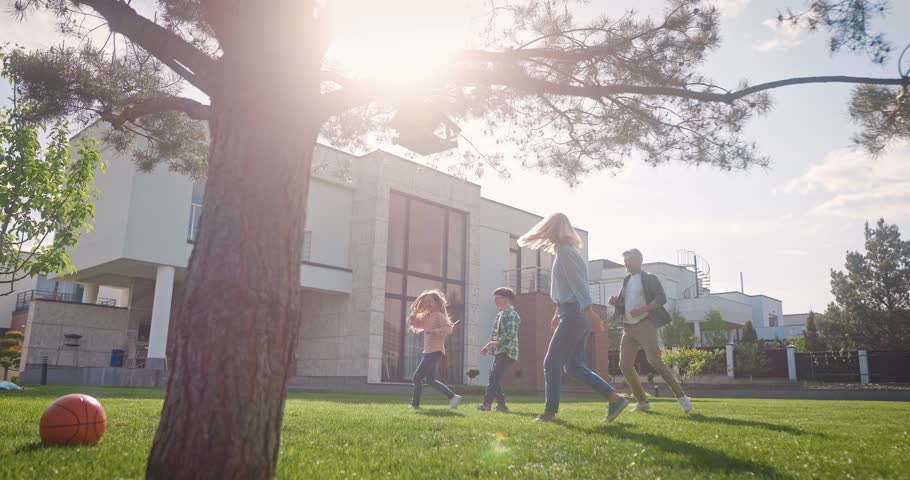 View of lovely Caucasian family running around next to tree in their backyard. Playing catch with their children in front of their new home. Spending time together as beloved and happy family.