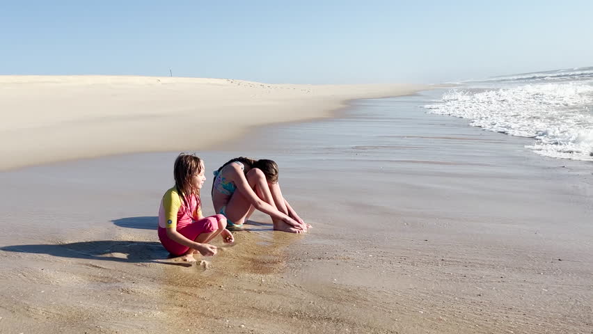 Video of two girls or sisters sitting on the beach, enjoying the water, with one in a pink swimsuit and the other in a colorful swimsuit. Children are playing on the beach