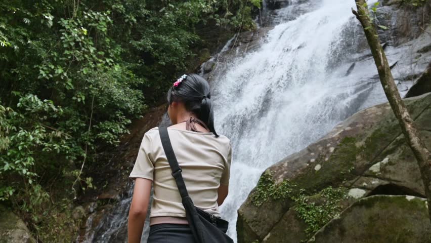 Rear view of relaxing pretty girl standing in front of stunning waterfall in tropical rainforest. Outdoor pursuit. Travel and Leisure activity. Ton Sung Waterfall, Phang Nga Province, Thailand.