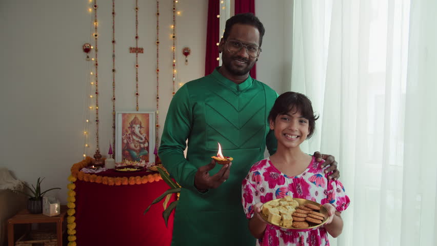 Medium long portrait of father and daughter of Indian ethnicity holding burning diya and plate of homemade cookies for Diwali at home