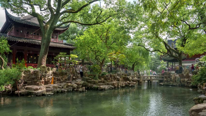 Visitors stroll along stone pathways at Yu Garden, surrounded by intricate wooden carvings, curved roofs, and a tranquil pond reflecting vibrant foliage.
