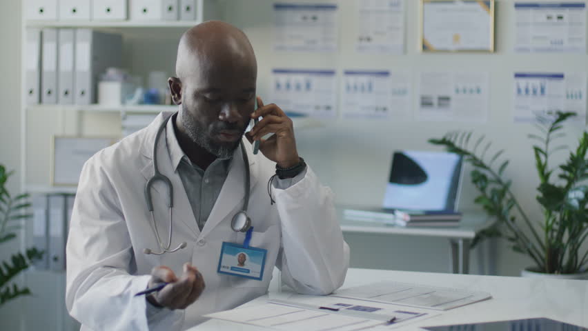 Black male doctor in medical coat sitting at desk in clinic, having conversation on mobile phone and taking notes on paper