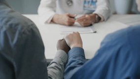 Middle-aged female doctor with grey hair smiling and talking to married couple holding hands during medical consultation in family clinic. Tilt-down, rack focus - Powered by Shutterstock - Get 15% off with code: PIKWIZARD15