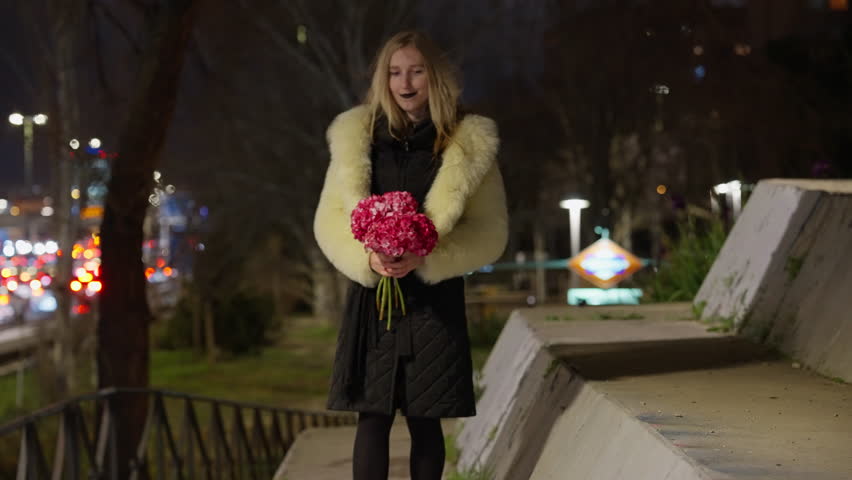 A cheerful young woman in a stylish outfit smiles brightly while holding a beautiful bouquet of flowers at night