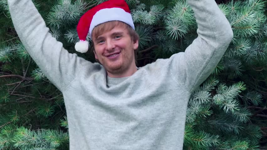 handsome man in front of christmas tree wearing santa claus hat holding gifts and rejoicing at christmas or new year. atmosphere, gifts, happiness