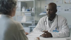 Grey-haired female patient handing medical records to Black male doctor, then answering his questions during medical consultation in clinic - Powered by Shutterstock - Get 15% off with code: PIKWIZARD15