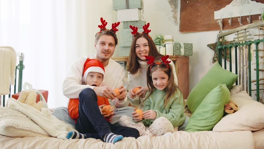 A cheerful family of four celebrates Christmas in cozy sweaters and reindeer antlers, watching TV and snacking on mandarins on a festive bed.