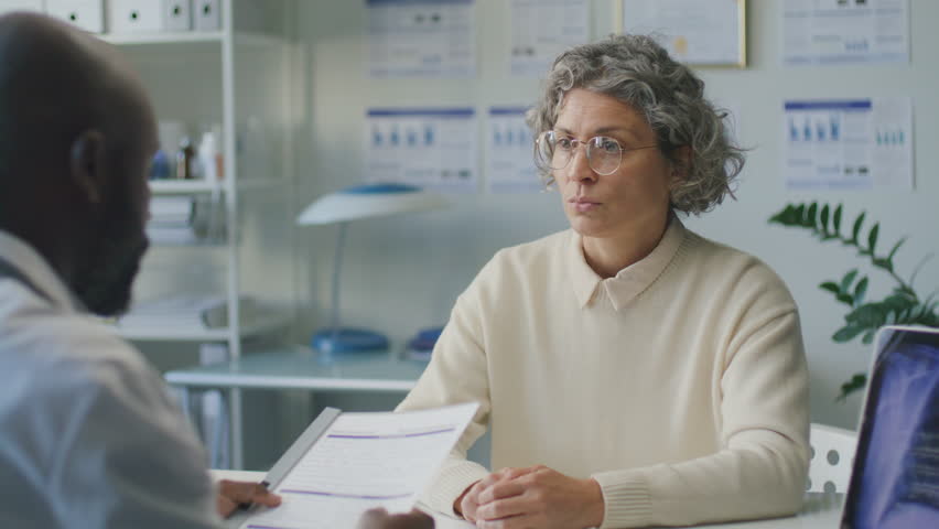 Middle-aged female patient with grey hair giving medical history to Black male doctor and talking to him during consultation in clinic