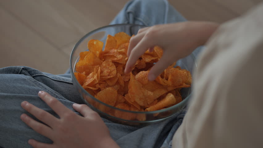 Girl Eating Chips From Bowl While Sitting on Floor in Living Room. Female Eating Unhealthy Potatoes Crisps. Snack on Junk Food. Child Eats Fast Food, Junk Food at Home Watching TV. Childhood Obesity.