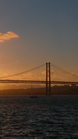 View of 25 de Abril Bridge famous tourist landmark of Lisbon connecting Lisboa and Almada on Setubal Peninsula over Tagus river with yacht silhouette on sunset and flying plane. Lisbon, Portugal