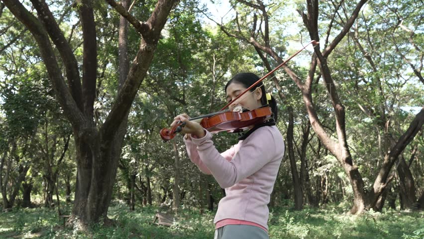 Young Asian woman enjoys playing the violin outdoors, sitting under a tree in the park. Surrounded by nature, she relaxes, practicing her music, embracing self-expression and international melodies.