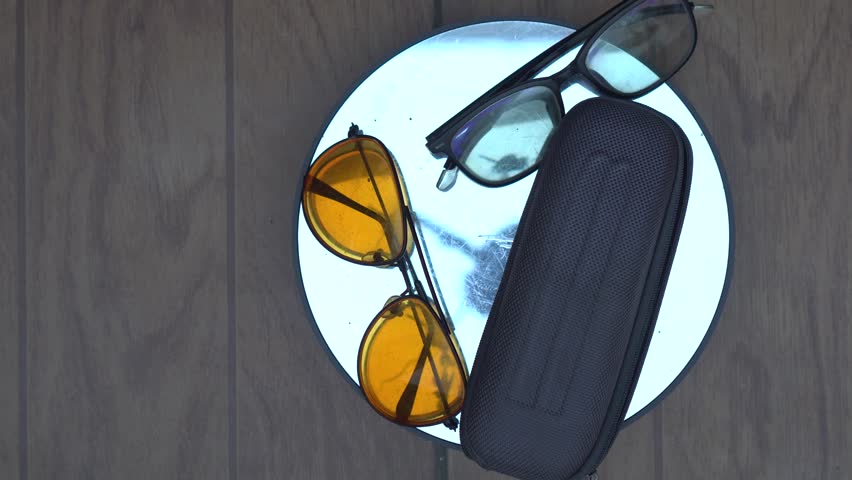 A pair of bright orange, round sunglasses sits on a sleek black case. The vintage look has a slight cat-eye shape, adding a fun retro touch. Next to them is a classic pair of black, 