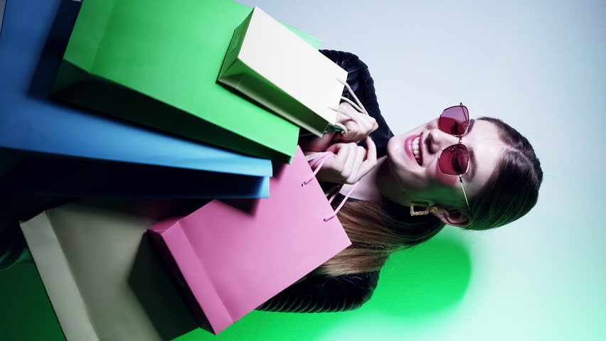 Vertical video. Shopping sale. Black Friday. Shopaholic lifestyle. Green neon light happy joyful glamour girl in glasses with bags enjoying purchase.