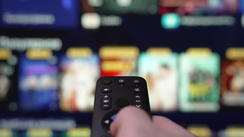 Person Browsing Streaming Services Using Remote Control. Hand Holding a Remote Control Browses Various Streaming Services On a Large Screen in a Comfortable Living Room Setting