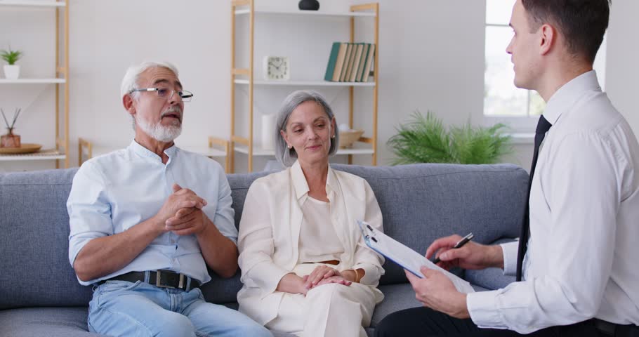 Senior gray haired couple sitting on sofa in living room, having consultation with financial agent about health insurance in retirement, carefully studying terms of investment contract before signing.